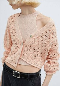 Meruňkový pletený svetr s otevřeným vzorem, se zapínáním na knoflík. Nositelný přes odpovídající žebrovaný top bez rukávů. Černý opasek zvýrazňuje pas.