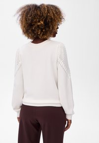 Crème gebreide trui met ballonsleeves, voorzien van een gestructureerd patroon op de schouders en een geribde zoom. Gecombineerd met donkerbruine broeken.