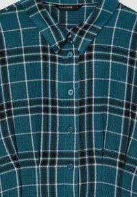 Camicia a quadri verde acqua con colletto, caratterizzata da un motivo a griglia in bianco e nero. Sei bottoni blu, tessuto testurizzato e due pieghe sul petto.