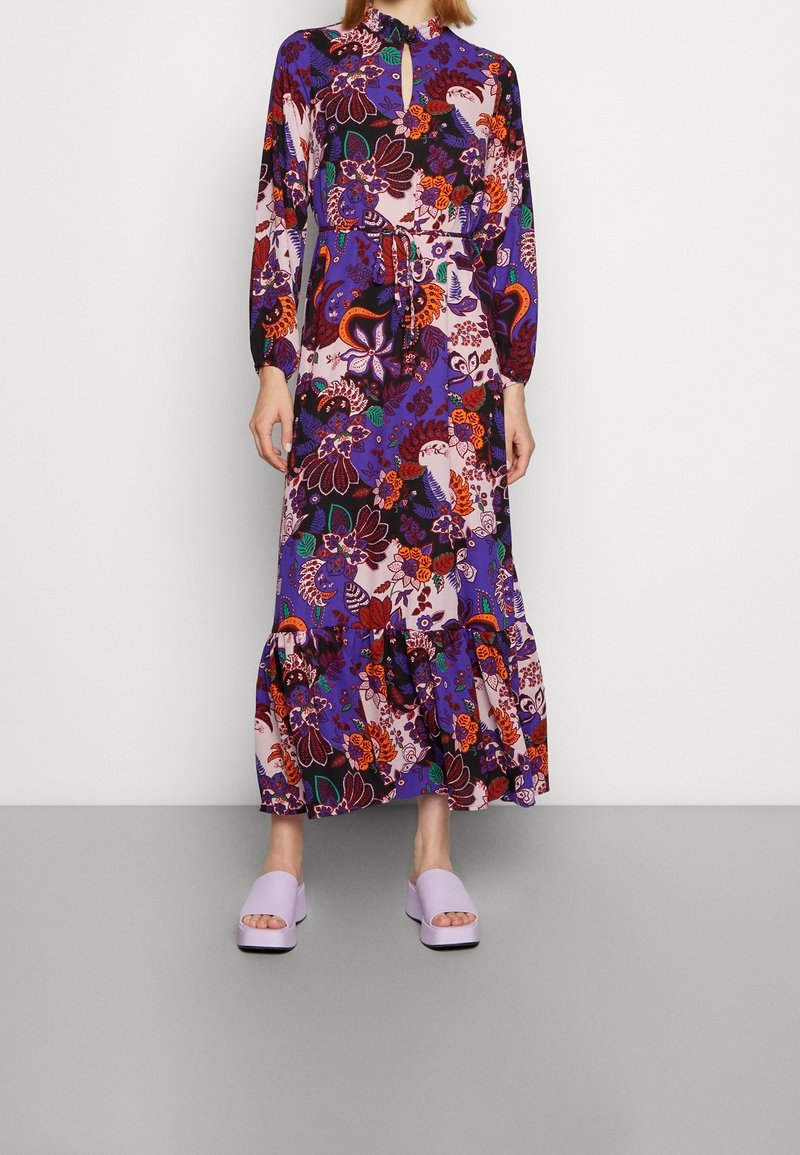 Femme portant une robe longue à manches longues, longueur cheville, avec des motifs floraux violets, orange et rouges, associée à des chaussures compensées ouvertes violettes claires.