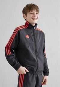 Mustat vetoketjulla varustetut urheilutakit, joissa on punaiset raidat ja logo. Ominaisuuksina korkea kaulus ja elastiset hihansuut. Kevyt kangas, jolla on sileä pinta.