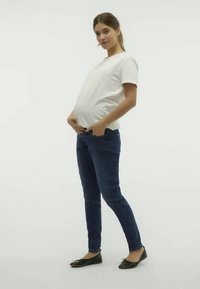 Bela kratka majica s kratkimi rokavi, oprijete kavbojke in črne balerinke, ki jih nosi noseča oseba. Drža poudarja zaobljen trebuh in sproščeno oblačilo.