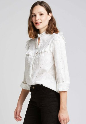 Blouse blanche brodée avec un col et des poignets à volants, ornée de motifs floraux et de petites découpes, associée à un pantalon noir.