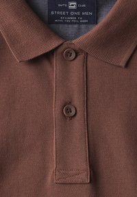 Camisa polo marrón con un tejido texturizado, dos botones marrones y un cuello clásico. Presenta una etiqueta con la marca "Street One Men".