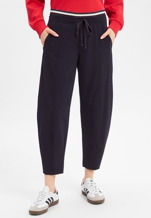 Personne portant un pantalon de jogging noir fuselé avec taille à cordon et des baskets blanches à rayures noires, mains dans les poches, haut rouge à manches longues.