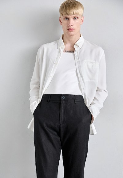 Mladá osoba s krátkými blond vlasy, oblečená v bílém rozepnutém košili přes bílý tílko a černé kalhoty, stojící u jednobarevné stěny.