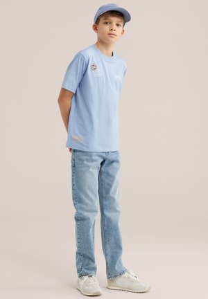 Fant, ki stoji z rokami za hrbtom, nosi svetlo modro kapo, svetlo modro majico z našitki, svetlo modre kavbojke in bele superge.