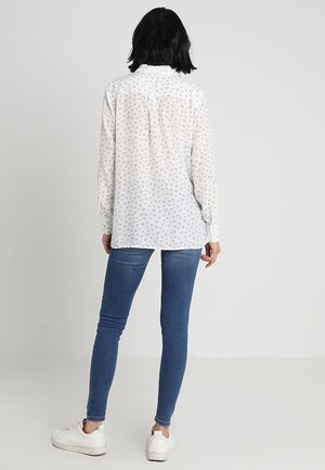 Femme aux cheveux courts noirs portant une chemise blanche à manches longues avec de petits motifs en forme de cœur, un jean skinny bleu et des baskets blanches, debout de dos.