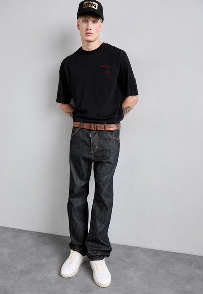 Jeune homme portant une casquette noire, un t-shirt graphique noir, un jean sombre avec une ceinture marron et des baskets blanches, se tenant devant un mur gris uni.