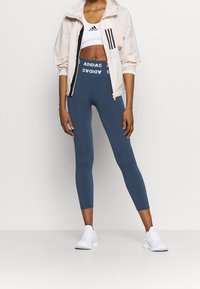 Chaqueta blanca recortada con cremallera negra y tres rayas, combinada con leggins azul marino que presentan el logo "ADIDAS" en la cintura y zapatillas blancas.