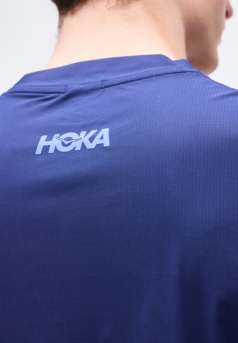Vista posteriore di una persona che indossa una maglietta sportiva blu con trama, con il logo "HOKA" centrato sotto la linea del collo.