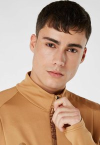 Beige zip-jakke lavet af glat stof, med høj krave og et mønstret lynlåsdetaile. Nærbillede af overkroppen og ansigtet.