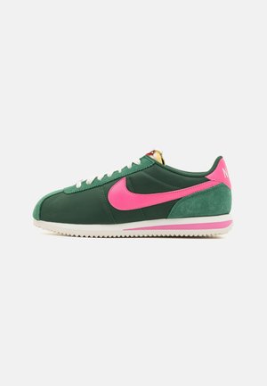 Lage groene sneaker met witte veters, roze Nike-swoosh en hielaccent, witte zool met roze streep, zijaanzicht.