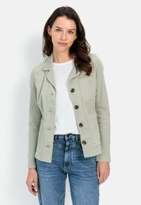 Lichtgroene jas met vijf knopen aan de voorkant, twee borstzakken en lange mouwen, gecombineerd met een wit shirt en spijkerbroek.