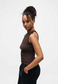 Jeune femme aux cheveux bouclés attachés en chignon haut, portant un haut marron sans manches et un pantalon noir, debout avec une main dans la poche.