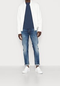 Giacca bomber bianca sopra una maglietta navy, abbinata a jeans blu chiaro e sneakers bianche. I jeans sono risvoltati alle caviglie.