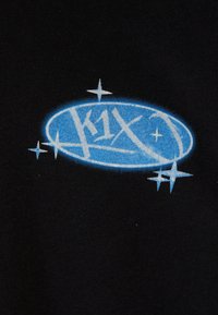Melna auduma virsma ar zilu ovālu logo, kurā baltā krāsā ir uzraksts "K1X" un ap to ir novietoti zvaigžņu apļu akcenti, demonstrējot gludu tekstūru.