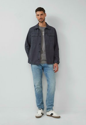 Grau gefütterte Jacke mit Knopfriegel, zwei Brusttaschen und einem schlichten grauen T-Shirt, kombiniert mit hellblauen Jeans und weißen Sneakers mit dunklen Akzenten.