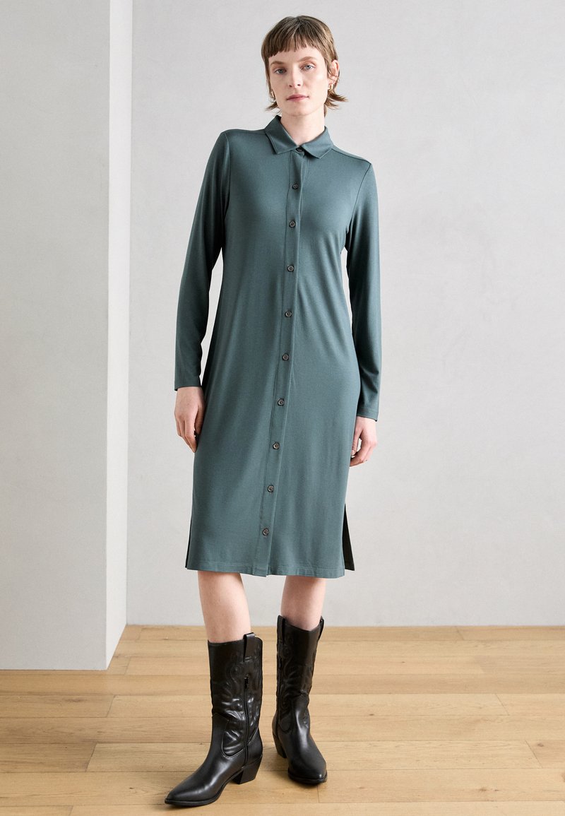 Robe verte à manches longues, boutonnée, en tissu doux, avec un col, des fentes latérales et des bottes de cow-boy noires jusqu'au genou.