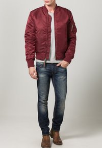 Muž v burgundské bomber bundě, šedém svetru, modrých džínách a hnědých botách, stojící s jednou rukou v kapse.