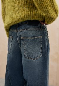 Jeans de ganga azul de corte solto com um acabamento desgastado, apresentando dois bolsos traseiros e costuras em destaque, combinados com um suéter amarelo texturizado.