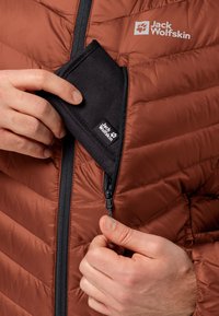 Veste marron gonflante avec une poche zippée, dotée d'une pochette en tissu noir. Le zip est noir et la veste a une texture matelassée.