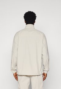 Giacca leggera di un beige chiaro, con colletto alto, maniche lunghe, polsini elasticizzati e una trama morbida. Presenta una vestibilità comoda e un design minimale.