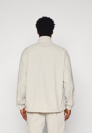 Giacca leggera di un beige chiaro, con colletto alto, maniche lunghe, polsini elasticizzati e una trama morbida. Presenta una vestibilità comoda e un design minimale.