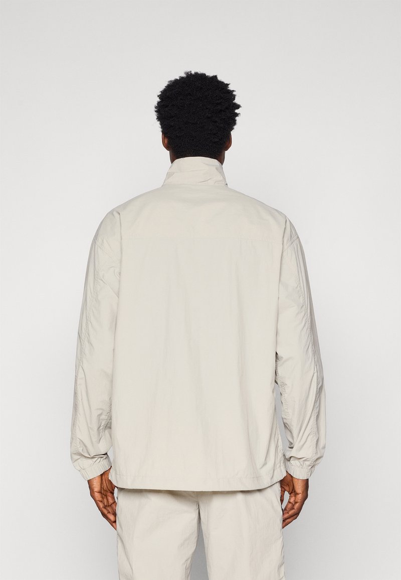 Giacca leggera di un beige chiaro, con colletto alto, maniche lunghe, polsini elasticizzati e una trama morbida. Presenta una vestibilità comoda e un design minimale.