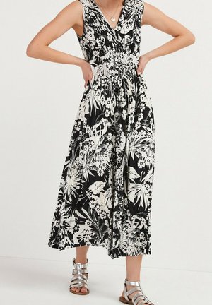 Femme portant une robe midi sans manches noire et blanche à imprimé floral avec un col en V et des sandales argentées à brides, debout les mains sur les hanches.