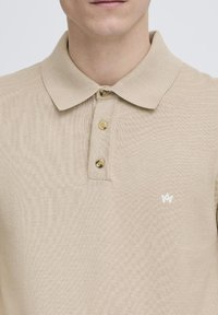Cămașă polo bej, fabricată dintr-un material tricotat texturat, având un guler clasic, placket cu trei butoane și un mic logo alb pe piept.