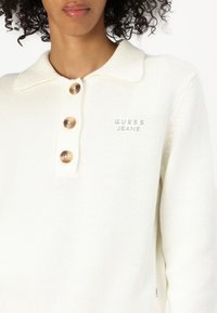 Biely pletený sveter s golierovým výstrihom, ktorý má tri veľké gombíky a text "GUESS JEANS" v sivej vyšívke na hrudi.