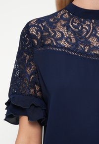 Námornícka blúzka s čipkovým zdobením a zložitými kvetinovými vzormi, volánovými rukávmi a hladkým textilným telom.