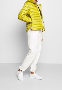 Kirkas keltainen puffatakki hupulla, jossa on tikattu muotoilu ja vetoketjusuljenta. Yhdistetty valkoisiin collegehousuihin ja valkoisiin tennareihin.