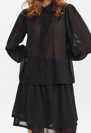 Femme portant un chemisier noir transparent à pois avec de longues manches bouffantes et une jupe assortie à volants.