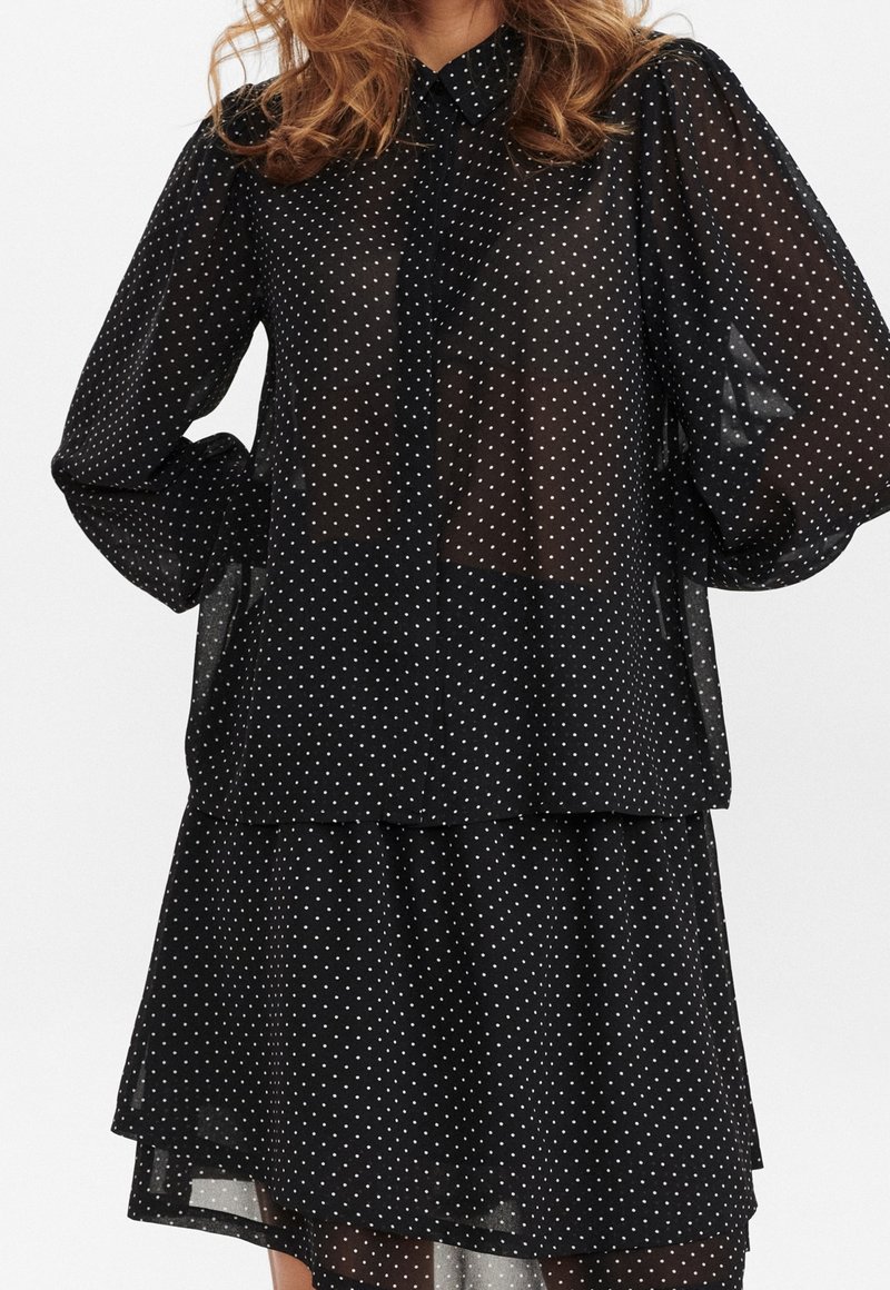 Femme portant un chemisier noir transparent à pois avec de longues manches bouffantes et une jupe assortie à volants.
