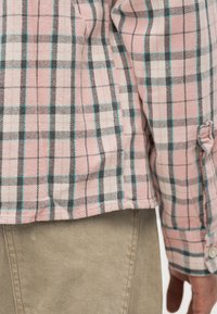 Ροζ, λευκό και μαύρο καρό μακρυμάνικο πουκάμισο με μανσέτες στα μανίκια, φορεμένο πάνω από μπεζ παντελόνι, με ένα ορατό χέρι να ακουμπάει στη πλευρά.