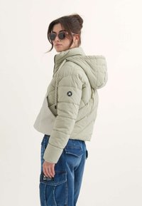 Una mujer con gafas de sol se gira ligeramente, vestida con una chaqueta acolchada verde clara y pantalones cargo de mezclilla azules, contra un fondo simple.