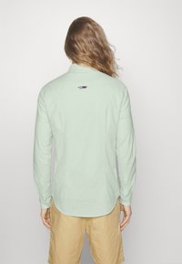 Mintgrön skjorta med knappar, lång ärm, rund nederkant och en liten triangel-logotypdetalj på övre ryggen. Tyget verkar slätt.