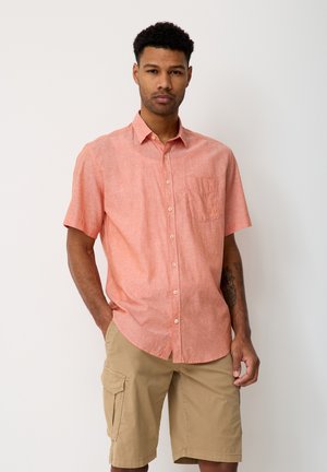 Man wearing coral short-sleeve button-up shirt and beige cargo shorts, standing with one hand in pocket against plain white background.