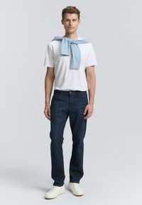 Joven vestido con una camiseta blanca, jeans azules, zapatillas blancas, y un suéter azul claro sobre los hombros.