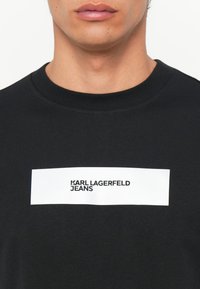 Černé bavlněné tričko s kulatým výstřihem, se bílým obdélníkovým grafickým prvkem, na kterém je tučně napsáno "KARL LAGERFELD JEANS" černými písmeny.