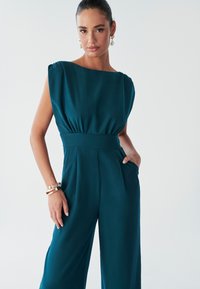 A woman poses confidently in a teal jumpsuit with a structured neckline and wide legs, accessorized with elegant earrings and bracelets.