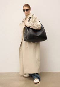 Sac à main en cuir noir avec une texture lisse, de grande taille et des accents en métal doré. Le modèle porte un trench beige et des chaussures blanches.
