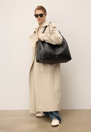 Sort lædertaske med en glat tekstur, stor form og guldhardware. Modellen bærer en beige trenchcoat og hvide sko.