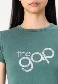 Zelené bavlnené tričko s krátkymi rukávmi a okrúhlim výstrihom; má veľké biele logo s nápisom "the gap" v štýlovom písme.