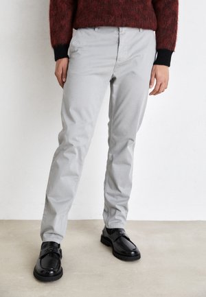 Persoon draagt lichtgrijze rechte broek met zwarte leren loafers en een donkerrode trui met zwarte manchetten, staand tegen een witte muur.