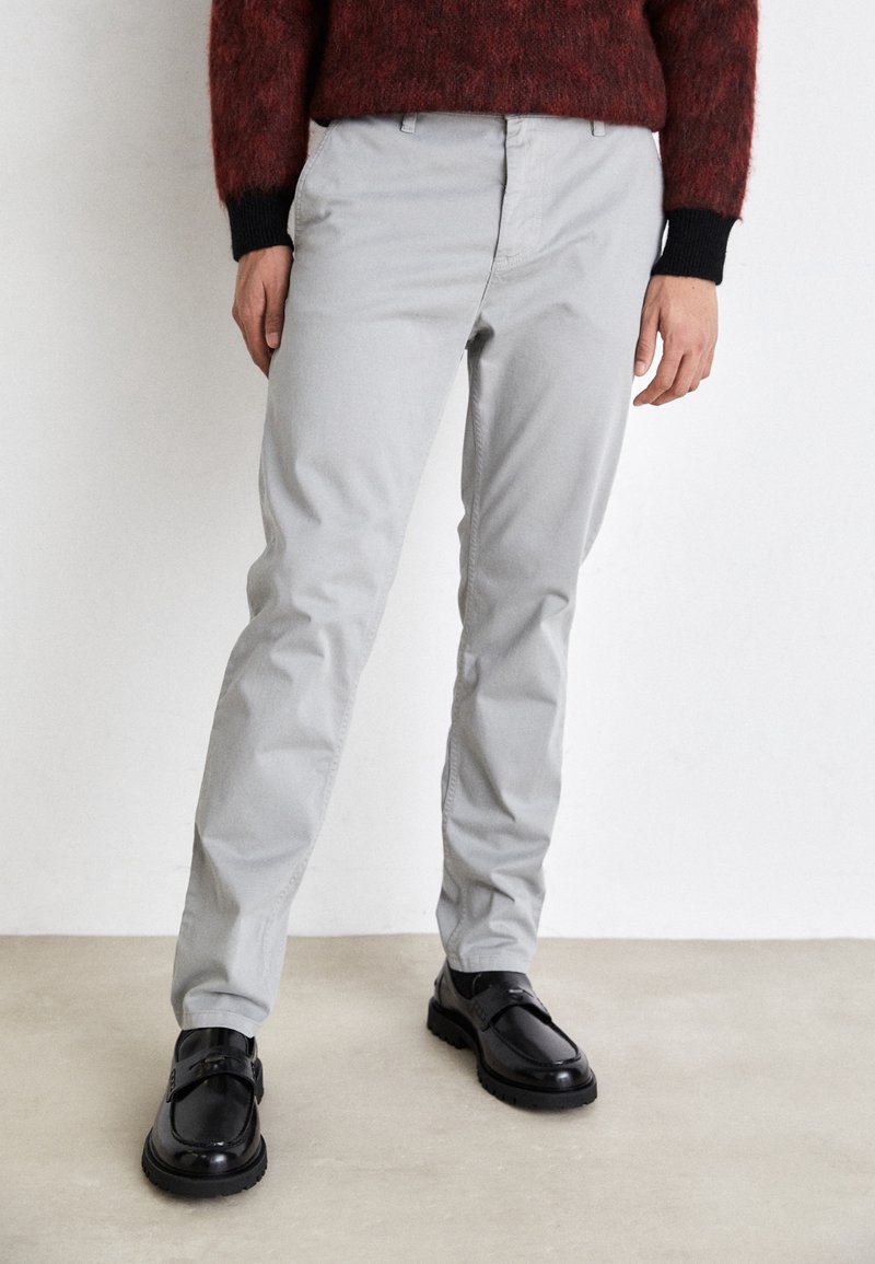 Personne portant un pantalon gris clair à jambes droites avec des mocassins en cuir noir et un pull rouge foncé avec des poignets noirs, debout contre un mur blanc.
