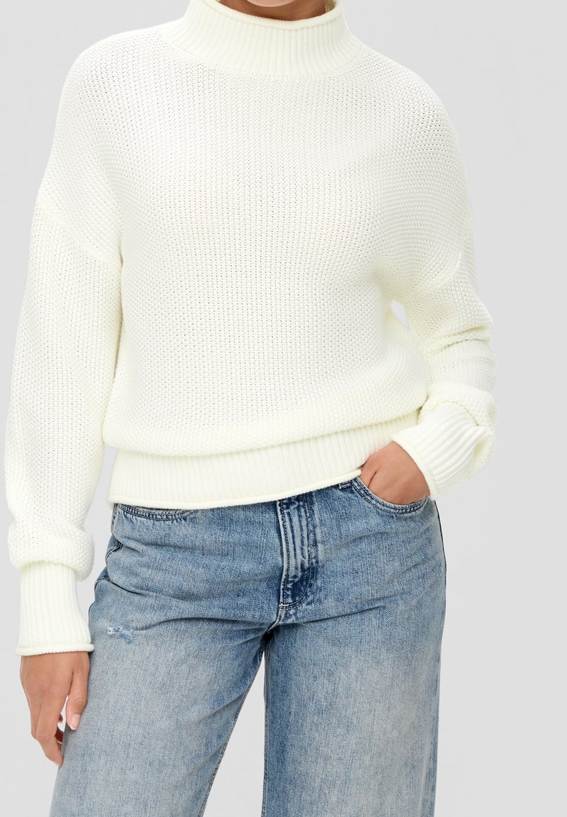 Biały sweter dziany z wysokim kołnierzem, luźnym krojem i ściągaczami, w połączeniu z jasnoniebieskimi podartymi jeansami.