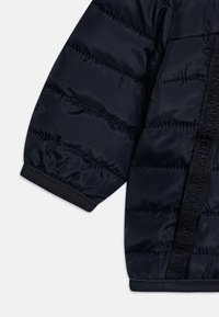 Chaqueta acolchada azul marino con un tejido texturizado, puños elásticos y un panel lateral con un detalle de cinta tejida con la marca.